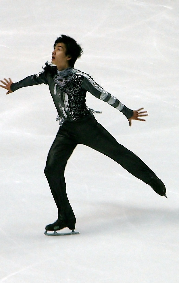 Ein männlicher Eiskunstläufer in einem schwarzen Kleid gleitet über ein Eisstadion und führt eine Routine mit synchronisierten Arm- und Beinbewegungen aus.