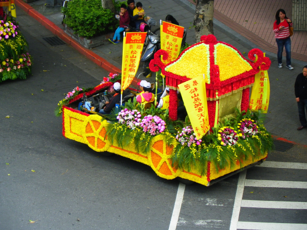 Ein Festwagen, der mit Blumen und Bannern geschmückt ist und Reiter trägt, mit Zuschauern auf der rechten Seite und Grün im Hintergrund.