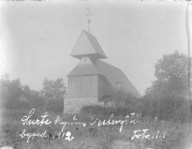 Schwarzes und weißes Foto der Kirche des Heiligen Grabes, eine Kirche mit einem Kirchturm, umgeben von Bäumen und Pflanzen unter einem bewölkten Himmel.