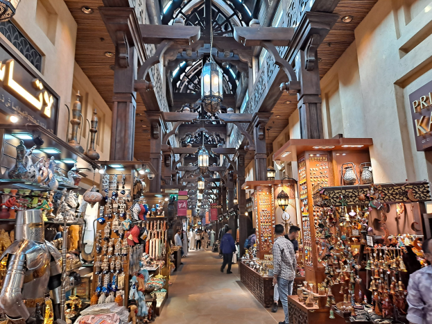 Ein geschäftiges Dubai-Bazar mit Menschen, die zwischen Regalen mit Waren hin und her gehen, Wänden mit Texttafeln und einer Decke mit warmem Licht.