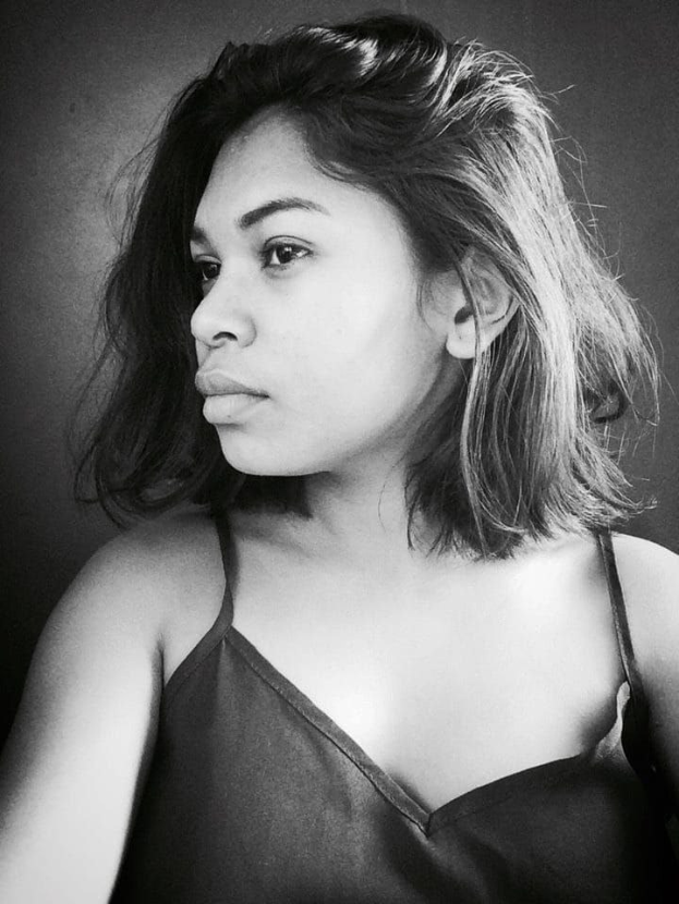 Eine selbstbewusste Frau in einem schwarzen Kleid posiert in einem Schwarz-Weiß-Foto, ihr glatter Bob und das natürliche Make-up unterstreichen ihre selbstbewusste Haltung an der Wand.