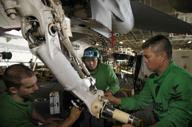 Drei Männer in grünen T-Shirts arbeiten an einem Flugzeug in einer Halle, einer trägt einen Helm, mit Equipment und Deckenbeleuchtung im Hintergrund.