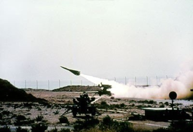 Eine Rakete startet von einem Militärfahrzeug in einer Wüstenlandschaft mit Vegetation, einem Zaun und Himmel im Hintergrund, mit Text oben.