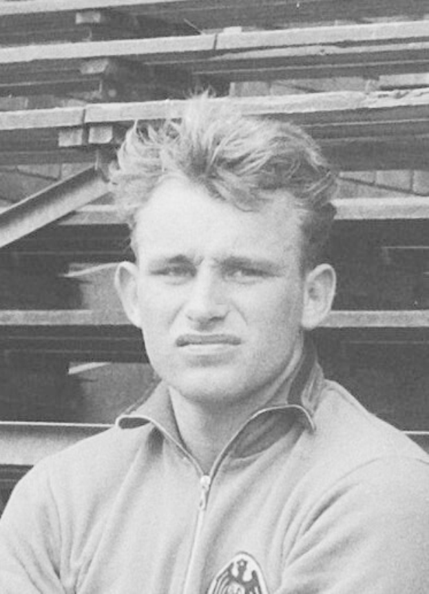 Schwarzes und weißes Foto eines Mannes in einer Jacke, der vor einem Haufen Holzplanken steht, der angeblich der legendäre deutsche Fußballspieler ist.