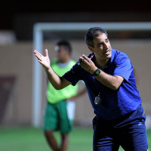 Ein Mann in einem blauen T-Shirt und Hose steht auf einem Fußballfeld und hält ein Mikrofon in der Hand, während er als Trainer gestikuliert. Im Hintergrund ist eine Person in einem grünen Kleid und ein Tor zu sehen.