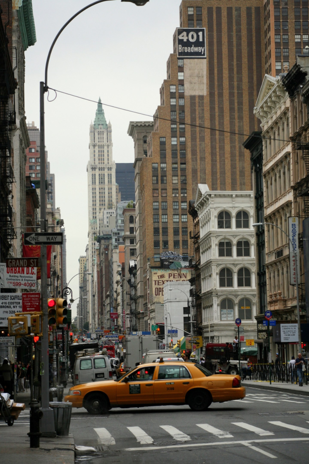Ein gelbes Taxi fährt auf einer Stadtstraße mit hohen Gebäuden, Fußgängern auf dem Gehweg, Verkehrsampeln und Textanzeigen, unter einem sichtbaren Himmel.