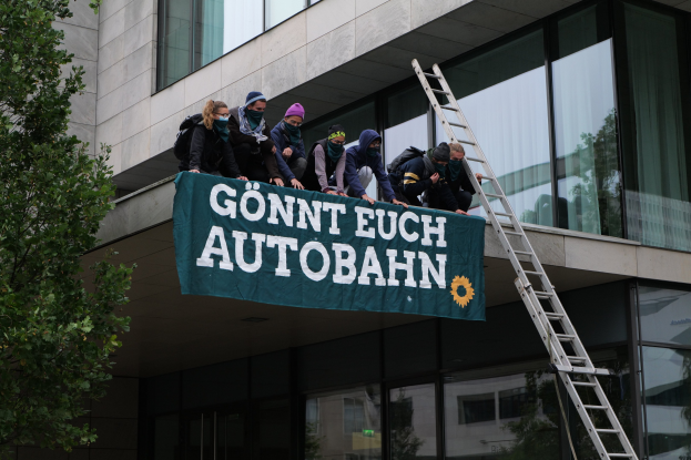 Eine Gruppe von Menschen auf einem Gebäude mit einer Fahne, mit einem Baum auf der linken Seite und Glasfenstern im Hintergrund, wahrscheinlich protestierend für die deutsche Regierung.