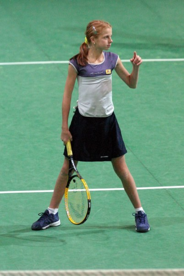 Eine junge Frau in einem weißen und blauen Tennis-Outfit steht auf einem Tennisplatz und hält einen Schläger in der Hand, ihr Haar ist zu einem Pferdeschwanz gebunden und sie hat einen entschlossenen Gesichtsausdruck.