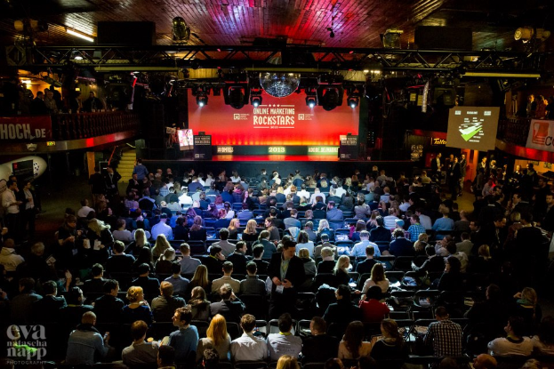 Eine große Menschenmenge sitzt auf Stühlen vor einer Bühne mit Bildschirmen und Plakaten, die von Scheinwerfern beleuchtet wird und von Geländern umgeben ist, und wartet gespannt auf den Beginn eines Online-Marketing-Rockstars-Events, mit einem Wasserzeichen in der linken unteren Ecke.
