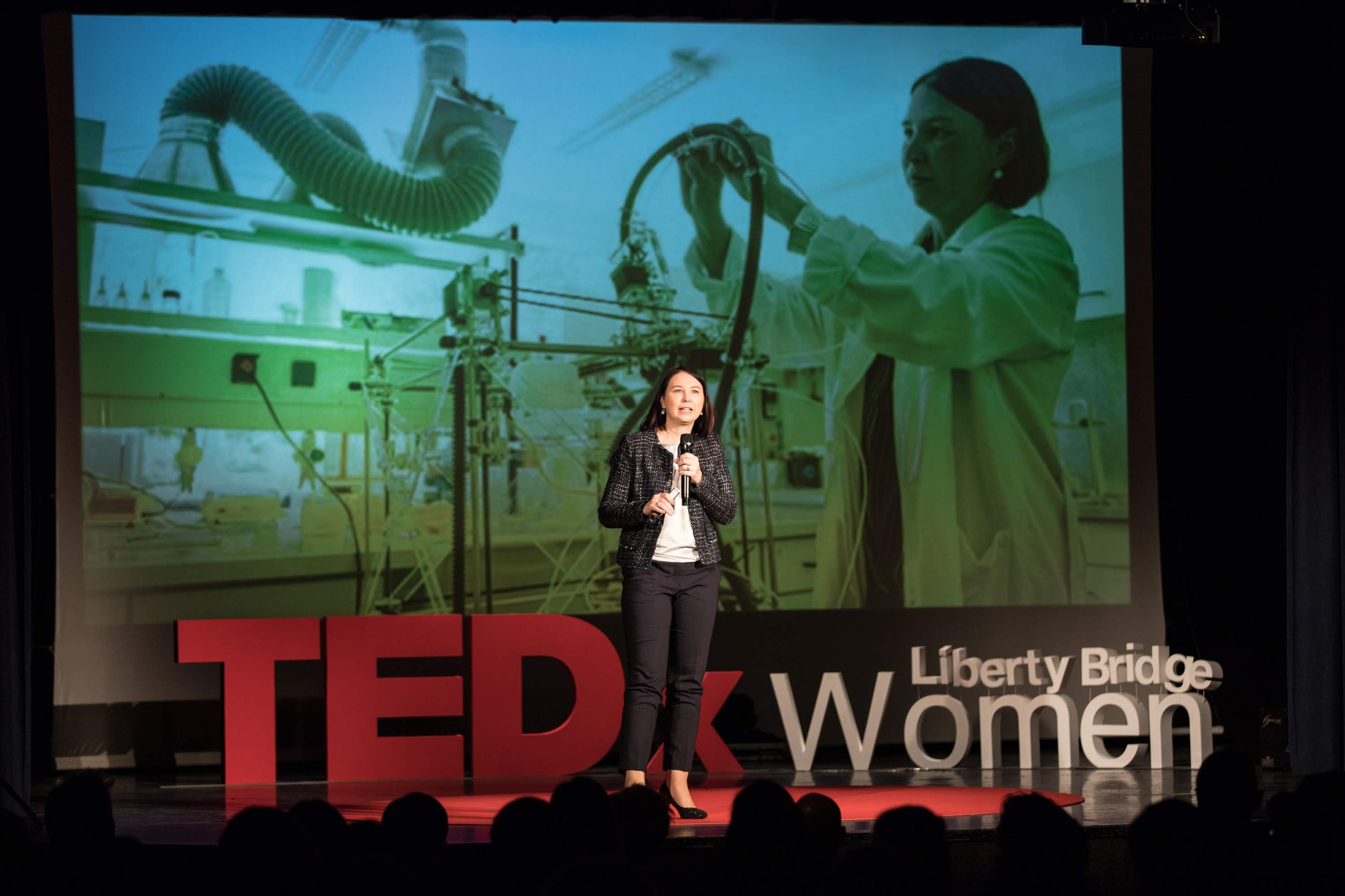 Frau auf einer Bühne mit einem Mikrofon, hinter ihr ein Display mit einem Bild einer Frau und Text zu einem TEDxWomen-Event.