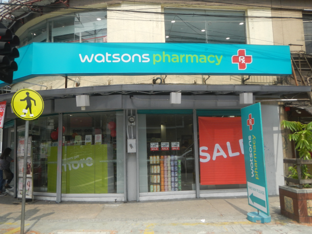 Ein Gebäude mit Glaswänden und -türen, ein Schild mit der Aufschrift "Watsons Pharmacy in New York City" auf einem Pfahl, eine Ampel mit einer Lampe, eine Gruppe von Menschen auf dem Gehweg, Topfpflanzen und ein bewölkter Himmel.