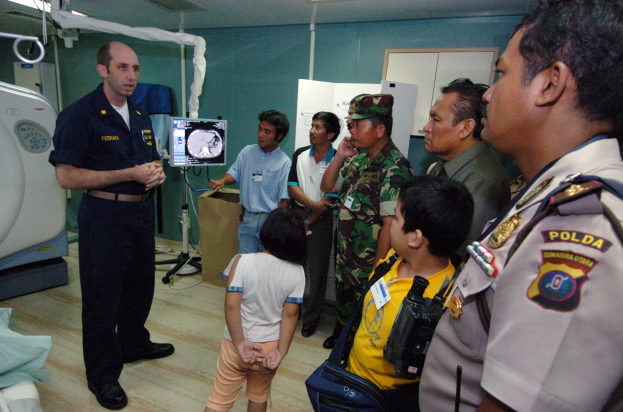 Eine Gruppe von Menschen steht in einem Krankenhauszimmer, ein Mann hält in der Mitte ein Mikrofon, links ein Bett mit einer Decke darauf und verschiedene medizinische Geräte, ein Röntgengerät, eine Tafel mit Text, eine Tür, eine Wand und eine Decke mit Lampen im Hintergrund.