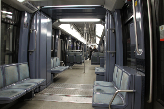 Innenseite eines U-Bahn-Wagens mit blauen Sitzen, Türen, Griffen, Stäben, Lichtern und einigen Passagieren in einer belebten Stadtkulisse.