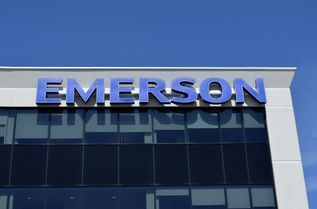 Das Emerson-Logo ist auf der Seite eines Gebäudes mit dem Text "Emerson investiert $1,5 Milliarden in den USA" darunter zu sehen, vor einem klaren blauen Himmel.