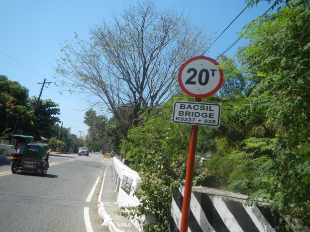 Eine Straße mit fahrenden Fahrzeugen, ein Tempolimit-Schild, eine Trennung, Bäume, Strommasten mit Drähten und einen klaren blauen Himmel.