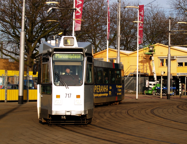 Ein weiß-blauer Tram auf einer Stadtstraße mit einer Person im Inneren, umgeben von Laternen, Bannern, einem Zaun, Fahrzeugen, Gebäuden, Bäumen und einem klaren blauen Himmel.