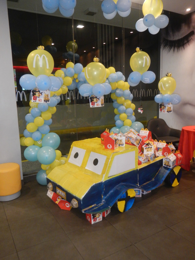 Eine festliche McDonald's Geburtstagsparty-Szene mit einem gelben und blauen Lastwagen, der mit Ballons geschmückt ist, einem rot gedeckten Tisch mit Gegenständen, Hockern und einer Glaswand im Hintergrund unter Deckenlampen.