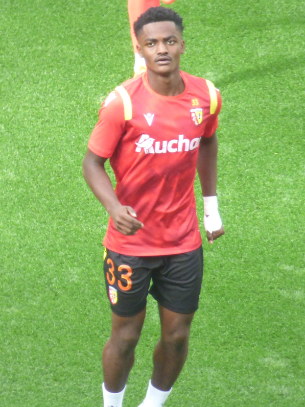 Emmanuel Adebayor in rotem Trikot und schwarzen Shorts auf einem Fußballfeld mit zwei Personen im Hintergrund.