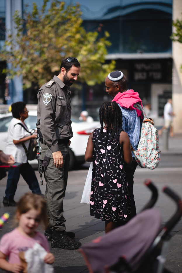 Ein Polizist spricht mit einer Gruppe von Menschen auf der Straße, mit einem Kinderwagen im Vordergrund und Fahrzeugen, Bäumen und einem Gebäude im Hintergrund.