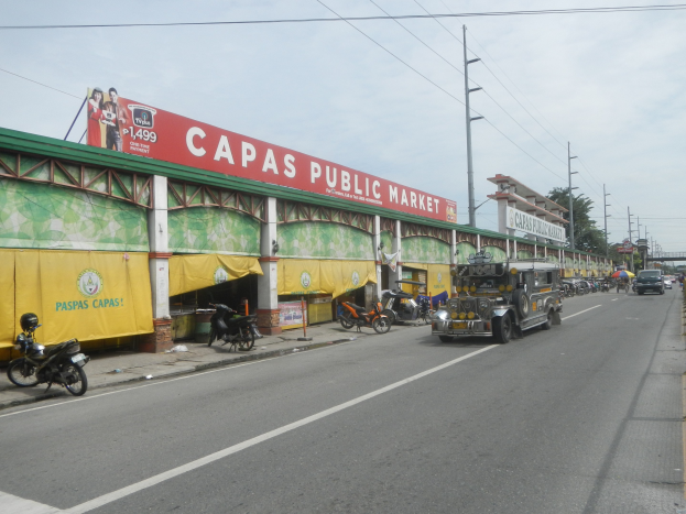 Eine belebte Straße mit Fahrzeugen, einem Gehweg, Strommasten, Gebäuden, Bäumen und einem bewölktem Himmel, mit einem Gebäude mit der Aufschrift "Capas Public Market" im Vordergrund.