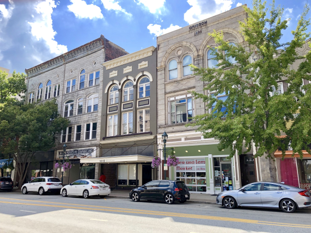 Eine Stadtstraße mit parkenden Autos, eine Mischung aus alten und neuen Gebäuden, Bäumen, Laternenmasten und einem bewölkten Himmel im Hintergrund.