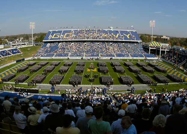 Eine große Menschenmenge in einem Stadion bei einem Fußballspiel, wobei einige stehen und andere sitzen, auf einem grünen Feld unter einem klaren blauen Himmel, mit Laternen, Bäumen und Gebäuden im Hintergrund.