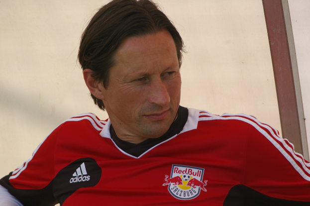 Ein professioneller Fußballspieler in einem roten und schwarzen T-Shirt sitzt auf einer Bank und schaut entschlossen.