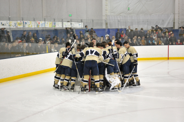 Gruppe von Hockeyspieler in dunklen Uniformen auf dem Eis während eines Spiels, mit Zuschauern im Hintergrund und Netz über der Wand.