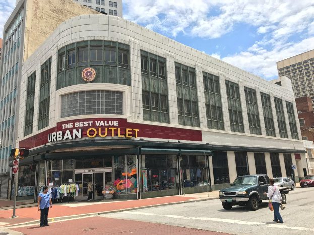 Eine belebte Stadtstraße mit Fahrzeugen auf der Straße und Fußgängern auf dem Gehweg, eingerahmt von hohen Gebäuden mit Glasfenstern und einem Schild mit der Aufschrift "The Best Value Urban Outlet" im Hintergrund unter einem bewölkten Himmel.