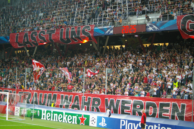 Ein Fußballspiel wird in einem Stadion mit einer großen Menge, grünem Rasen, einem Torpfosten links, Bannern auf dem Boden und Menschen mit Fahnen gespielt, mit Geländern, Tafeln, Lampen und anderen Gegenständen im Hintergrund.