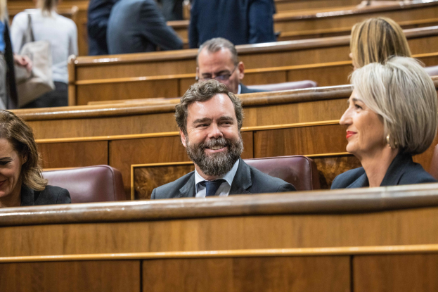 Gruppe von Menschen, die in einem Gerichtssaal sitzen, mit einem Mann in Anzug und Krawatte und zwei leger gekleideten Frauen, die nebeneinander sitzen, sowie Bänken im Hintergrund.