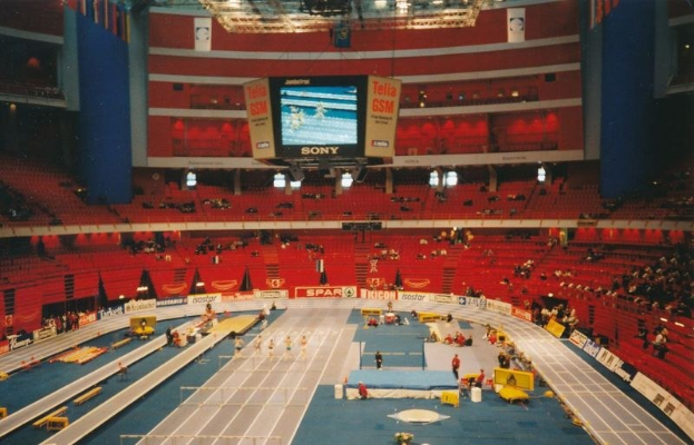 Großes Hallenstadion mit Zuschauern bei einer Leichtathletikveranstaltung, einschließlich sitzender und stehender Zuschauer, Texttafeln, Beleuchtung und einem Bildschirm oben.