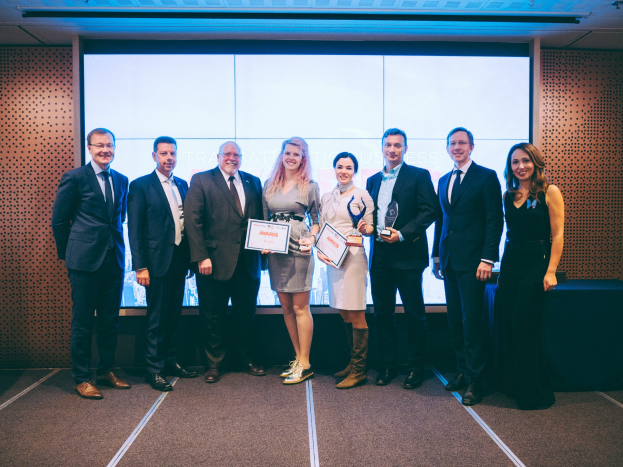 Eine Gruppe von lächelnden Menschen mit Urkunden, die vor einem Bildschirm mit einem Tisch und einem Wandobjekt im Hintergrund stehen, erkannt als Gewinner der Internationalen Business Awards 2018.