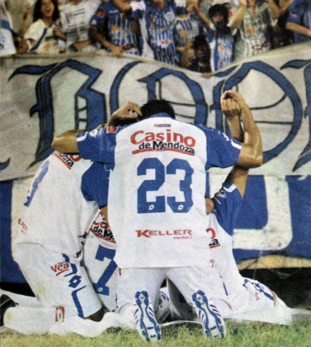 Gruppe von Fußballspielern, die auf dem Platz vor einer jubelnden Menge mit einem Banner im Hintergrund feiern.