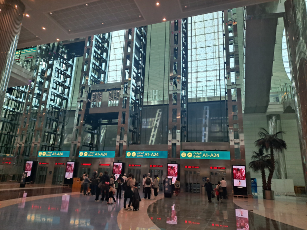 Menschen mit Gepäck durch eine Flughafenhalle gehen, umgeben von hohen Gebäuden, Bäumen, Texttafeln, Säulen und Deckenleuchten.