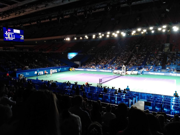 Ein Tennis-Match in einer großen Arena mit Zuschauern auf Stühlen, ein Netz teilt den Court, einige stehende Personen, ein Bildschirm mit Text auf der linken Seite und Überdachungsbeleuchtung.