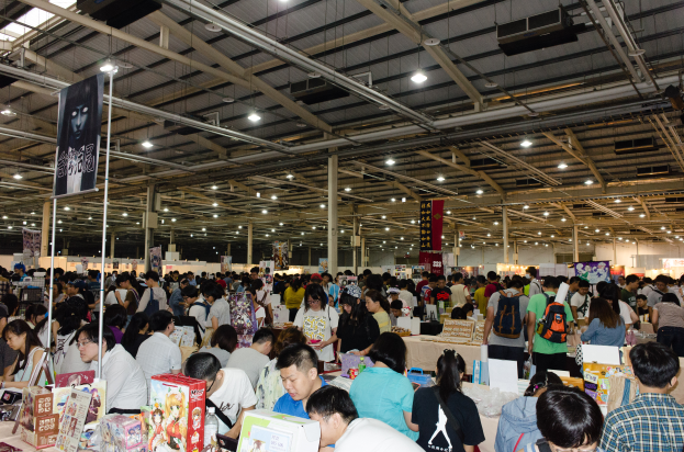 Eine große Gruppe von Menschen versammelt sich um Tische mit Büchern, Kartons und anderen Gegenständen auf einer Buchmesse, mit Schildern und Lichtern darüber.
