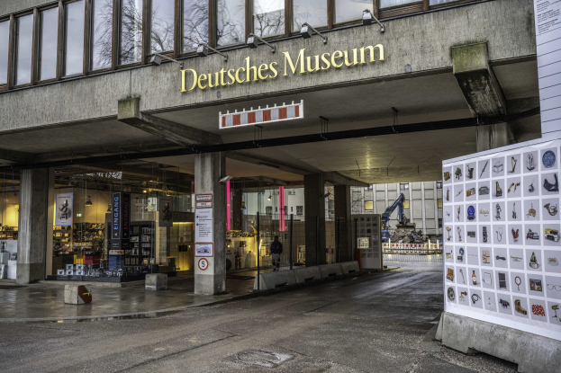 Außenansicht des Deutschen Museums in Berlin, das eine große Glasfassade mit Säulen, ein Namensschild, eine Anzeigetafel mit Bildern und Text sowie umliegende Strukturen, Bäume und Himmel zeigt.