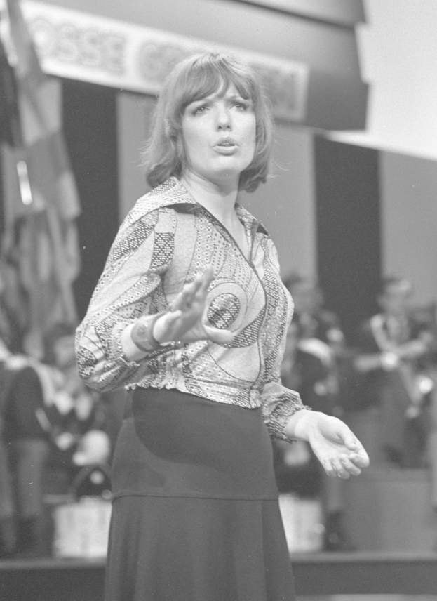 Eine Frau in einem Schwarz-Weiß-Foto steht auf einer Bühne, trägt ein weißes Hemd und einen schwarzen Rock, hat ihre Haare zu einem Dutt gebunden und einen ernsten Ausdruck im Gesicht, umgeben von einer Gruppe von Menschen und einer Wand im Hintergrund.