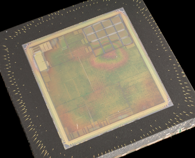 Nahaufnahme eines rechteckigen Halbleiterchips mit metallischem Glanz, dünnem Metallrahmen und kleinen Kantenlöchern auf schwarzem Hintergrund, mit leicht erhöhten Kanten.