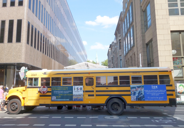 Ein gelber Schulbus fährt auf einer Straße mit hohen Gebäuden, Fußgängern auf dem linken Gehweg, Bäumen im Hintergrund und einem klaren blauen Himmel.