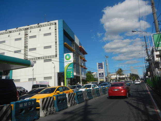 Eine belebte Stadtstraße mit zahlreichen Fahrzeugen, Absperrungen, Beschilderungen, Laternen, Strommasten, Bäumen und hohen Gebäuden.