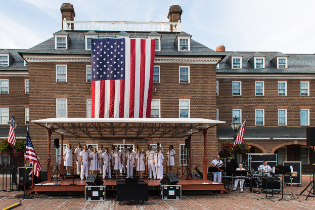 Eine Gruppe von Menschen auf einer Bühne am Mount Vernon, einige spielen Musikinstrumente, mit Fahnen, Lautsprechern, Pflanzen und einem bewölkten Himmel im Hintergrund.