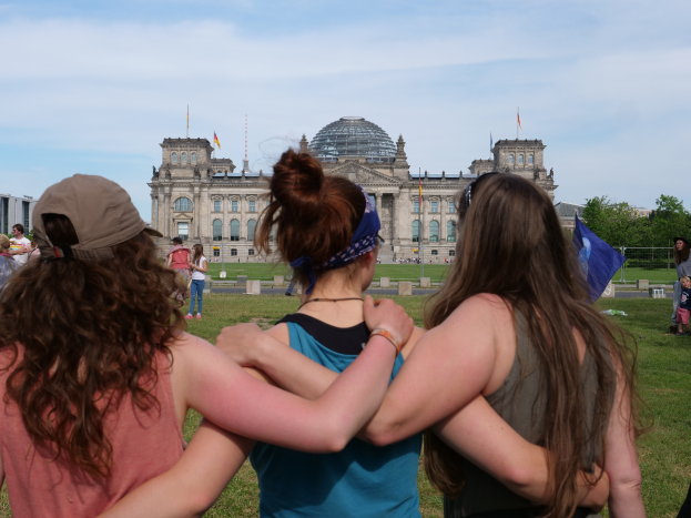 Drei Frauen in verschiedenen farbigen Kleidern, eine mit einer Mütze, stehen vor dem Reichstaggebäude in Berlin, Deutschland, mit Menschen, Fahnen, Bäumen, Wolken und Himmel im Hintergrund.