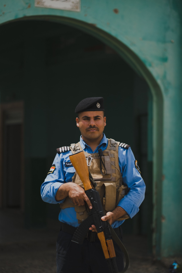 Polizeibeamter in Uniform mit Gewehr, vor einem Gebäude mit einem Tor und einer Tafel an der Wand stehend.