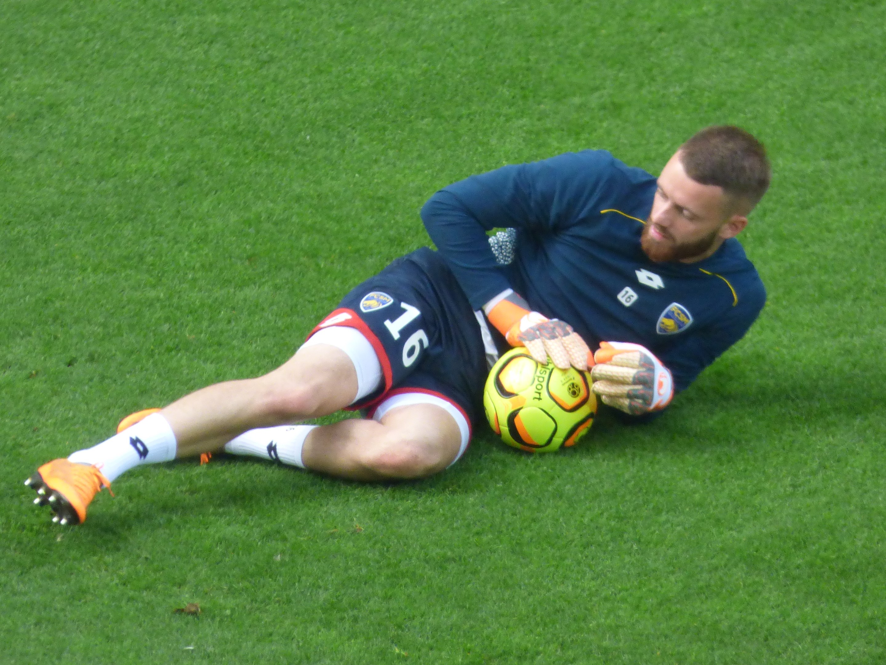 Ein Fußballspieler liegt auf dem Boden und hält einen Ball, trägt ein blaues T-Shirt, Shorts, Socken und Schuhe.