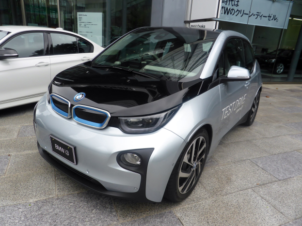 Ein silberner BMW i3 Elektroauto vor einem Gebäude in Tokio, Japan, mit Glaswänden und einer Texttafel im Hintergrund.