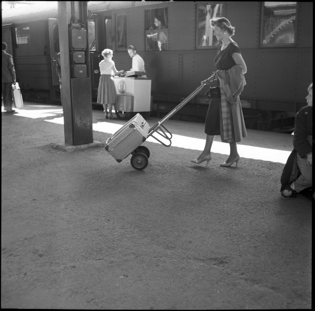 Eine Frau schiebt einen Kofferwagen mit einem Koffer an einem Bahnhof, umgeben von ein paar Menschen und einem Zug im Hintergrund, alles in Schwarz-Weiß dargestellt.