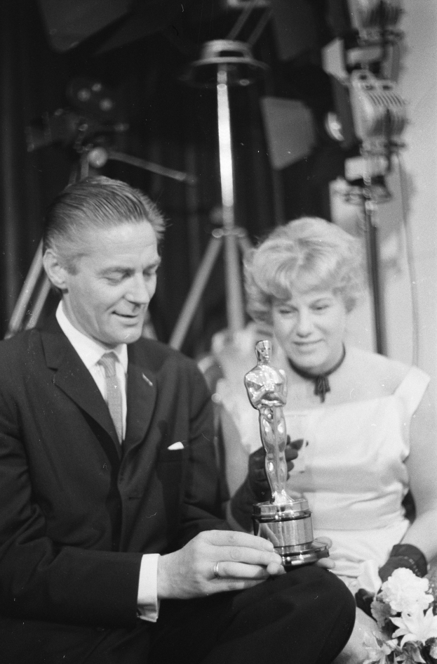 Ein Schwarz-Weiß- Bild eines formell gekleideten Mannes, der eine Oscar-Statue hält und eine Frau, die einen Blumenstrauß hält, nebeneinander sitzen, mit beleuchteten Lichtern im Hintergrund.