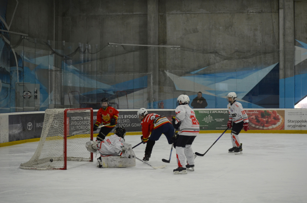 Gruppe von Menschen, die Eis hockey auf einem Eisplatz spielen, tragen Helme und halten Hockey-Schläger, mit einem Torpfosten auf der linken Seite und Bannern im Hintergrund.
